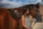 Young woman stands beside a saddled brown horse outdoors under a blue sky, looking calm.