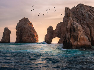 Rocher emblématique à Cabo San Lucas au coucher du soleil, avec des oiseaux volants et une mer calme, ciel aux teintes chaudes.