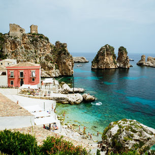 Coastal scene with turquoise water, rocky cliffs, and a red building. People sunbathe by the shore. Boats float in the clear sea.