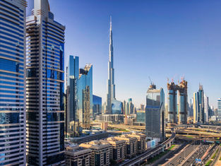 A panoramic daytime view of the Dubai skyline, featuring the iconic Burj Khalifa towering in the center amidst a dense cluster of modern skyscrapers and buildings under construction, with a network of busy multi-lane highways in the foreground under a clear blue sky.