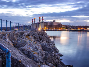 Industrial plant with lit chimneys overlooks a calm waterfront at dusk, surrounded by rocks and a blue-railed pathway under a cloudy sky.