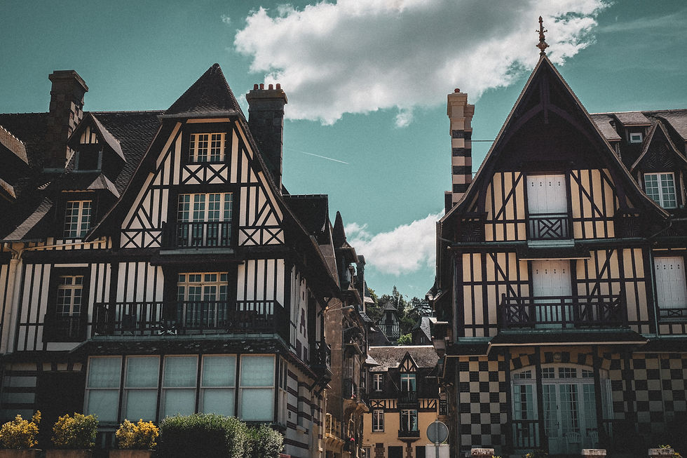 Half-timbered houses in northern France with steep roofs under a cloudy sky. A jet’s contrail is visible, adding a sense of motion to the tranquil scene.