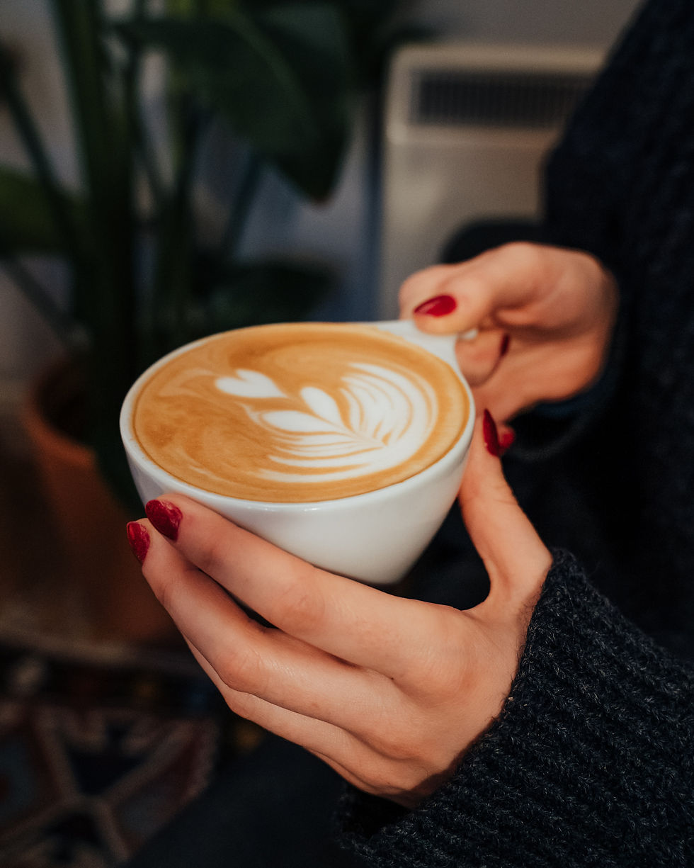 Hands with red nails hold a latte in a white cup, topped with leaf latte art, in a cozy café setting.