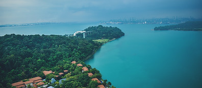 Aerial view of a lush green peninsula surrounded by blue water, with red-roofed buildings in the foreground and distant cityscape and port cranes under a cloudy sky.