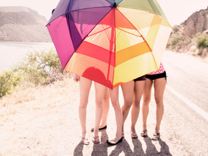 Four individuals stand under a large, colorful umbrella on a sunny roadside. Mountains and greenery in the background create a relaxed vibe.