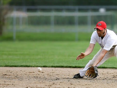 Baseball player in a white uniform and red cap crouches, reaching for a rolling ball on a grassy field, focused and determined.