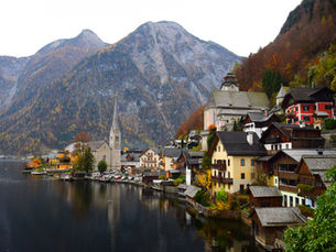 A picturesque view of Hallstatt, Austria, with traditional alpine houses nestled between a serene lake and majestic, tree-covered mountains under an overcast sky. A prominent church steeple rises in the center of the village.