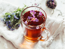 Clear glass mug with herbal tea and purple flowers on top, set on a white cloth. Fresh flowers and strainer in background. Peaceful mood.