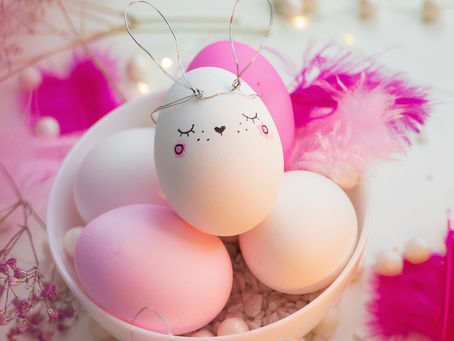 A charming Easter egg decorated with a whimsical bunny face and wire ears rests among pastel-colored eggs and vibrant pink feathers.