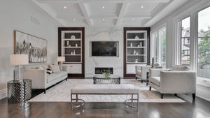 Elegant living room with a white rug and gray furniture. Large windows, a wall-mounted TV, artwork, and bookshelves create a cozy setting.