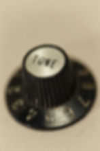 Close-up of a black, ridged guitar knob labeled "TONE," with surrounding numbers on a blurred beige background.