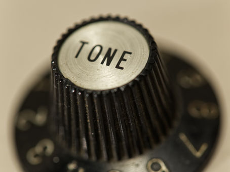 Close-up of a black, ridged guitar knob labeled "TONE," with surrounding numbers on a blurred beige background.