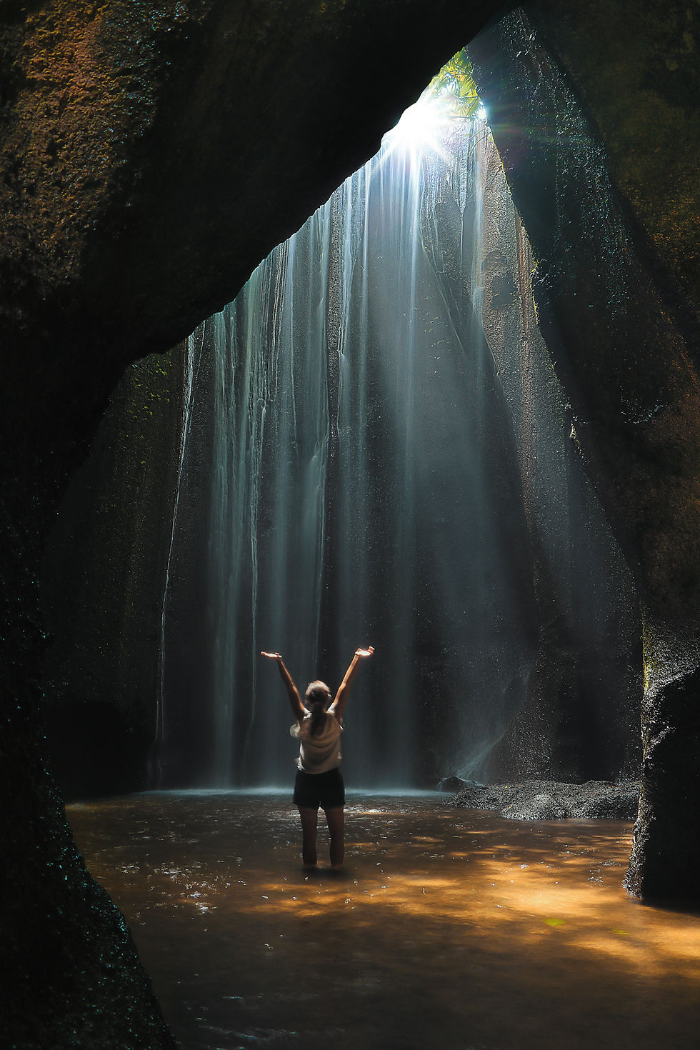 A person stands in awe, arms raised, beneath the enchanting light and cascading waters of Tukad Cepung Waterfall.