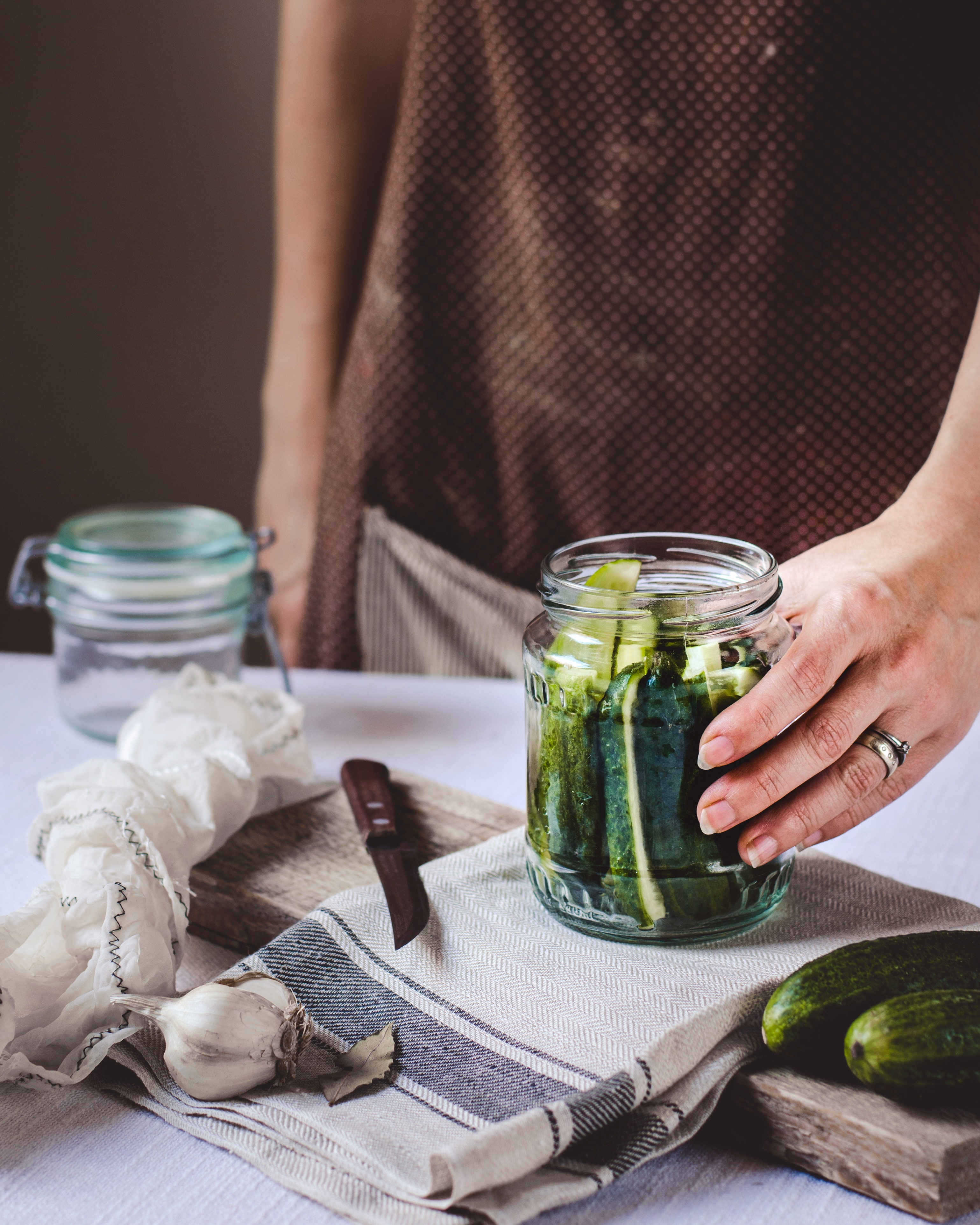 Hand placing pickles into a jar on a table with a knife, garlic, cloth, and empty jar in the background. Brown apron, rustic setting.