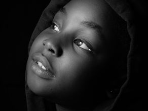 A child gazes upward in a black and white close-up, wearing a hooded garment. Soft lighting highlights a contemplative expression.