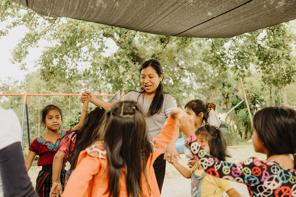 Woman and children play holding hands under a canopy. Background includes trees and a swing set. Kids wear colorful clothes; joyful mood.