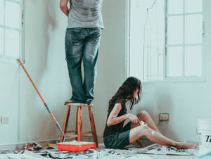 Person standing on stool painting a white wall, another sitting on the floor holding a brush. Paint supplies scattered around; sunlight streams in.