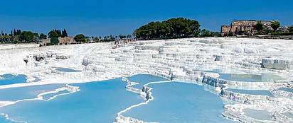 Terraced white mineral pools filled with turquoise water at Pamukkale, Turkey, with green trees and ancient ruins visible in the background under a clear blue sky.