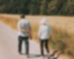 Two people walk away along a rural road beside tall grass, with sunlit trees in the background.
