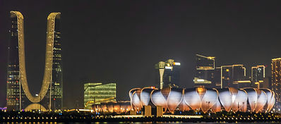 Night view of a modern cityscape featuring a tall, uniquely curved skyscraper on the left and a stadium with a lotus flower-inspired design, illuminated against the dark sky with other lit buildings in the background.