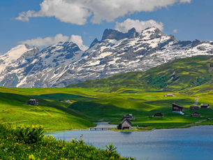 A stunning panoramic view of the Swiss Alps, featuring lush green, rolling hills dotted with traditional wooden chalets leading down to a serene, blue alpine lake. In the background, majestic, snow-capped mountains rise towards a blue sky with scattered white clouds.
