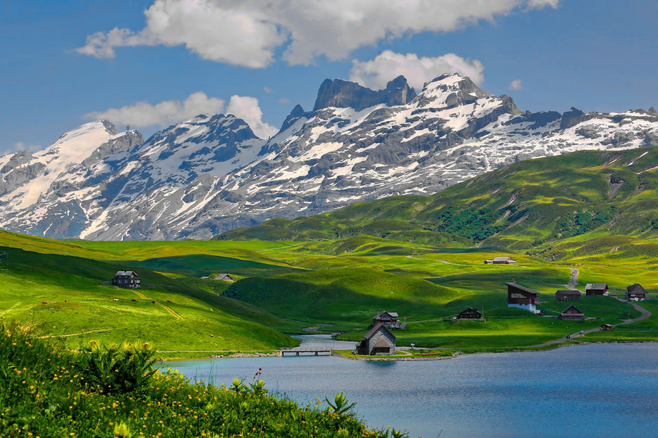 A stunning panoramic view of the Swiss Alps, featuring lush green, rolling hills dotted with traditional wooden chalets leading down to a serene, blue alpine lake. In the background, majestic, snow-capped mountains rise towards a blue sky with scattered white clouds.