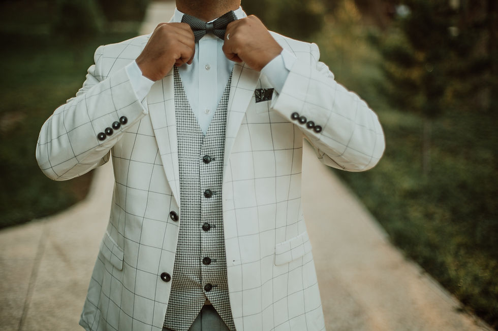 Man in white checkered suit adjusts bow tie outdoors on a path, wearing a patterned vest, with a blurred green background.