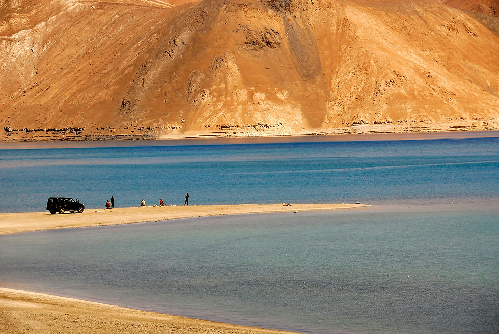 Jeep and people on a sandy beach by a serene lake, surrounded by barren mountains under a clear sky, conveying a peaceful mood.