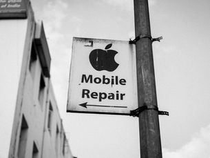 Sign with an apple icon reads "Mobile Repair" and points left. Mounted on a pole with a building in the background. Black and white image.