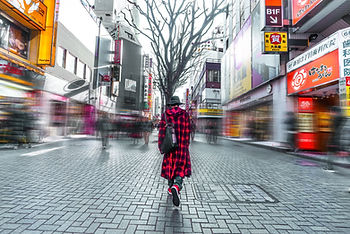 Person in a red plaid coat walks through a bustling city street with blurred crowds. Neon signs and buildings line the vibrant background.