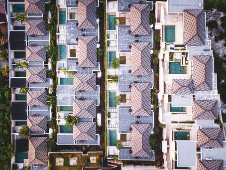Aerial view of a row of identical houses with brown roofs and pools. The geometric layout is surrounded by greenery, creating a serene mood.