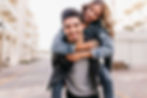 Smiling young couple on a sunny street, woman piggybacking on man in denim jackets, with bright buildings blurred behind them.