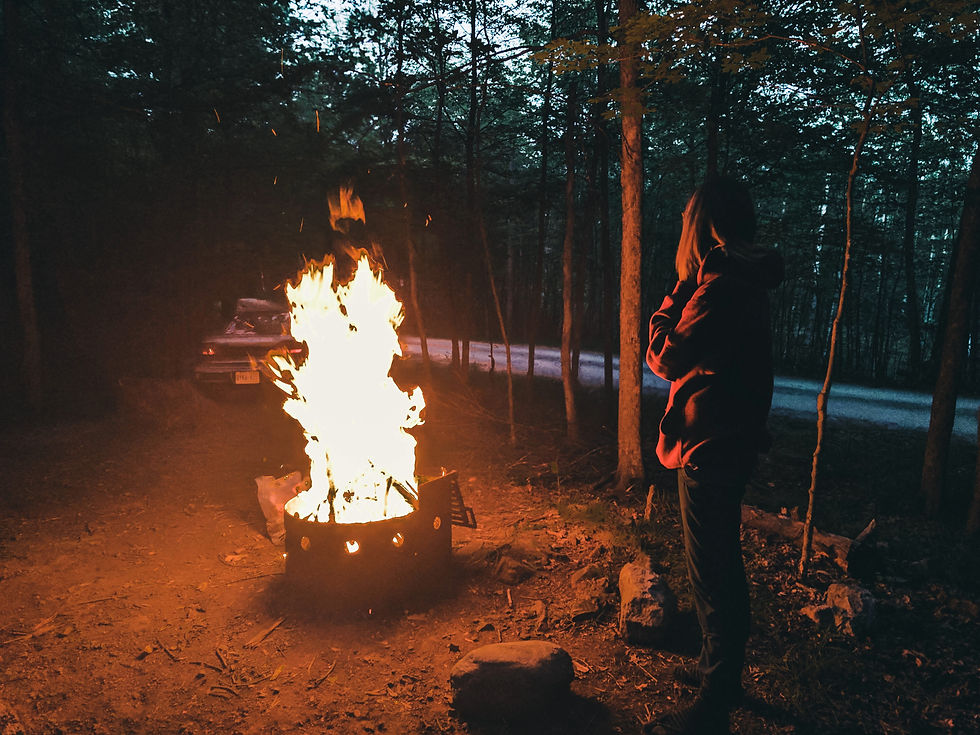 Person in hoodie stands near large campfire in forest. Car visible in background on road. Scene lit by fire's glow, evoking a cozy mood.