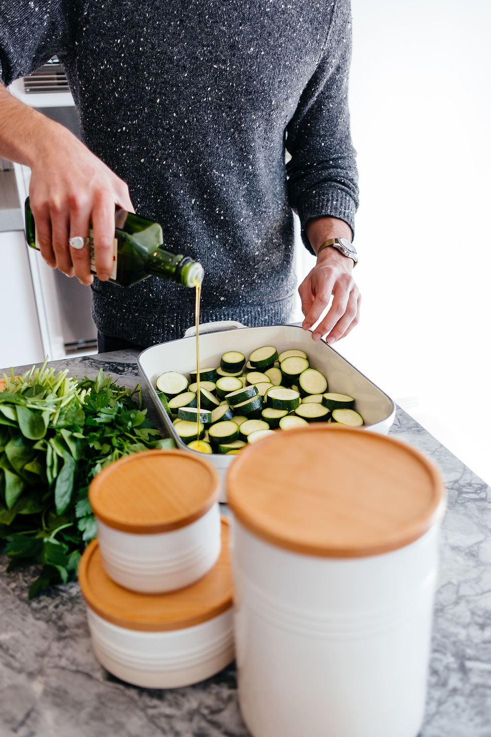 Person in a gray sweater pours olive oil over zucchini slices in a kitchen. Fresh greens and white canisters with wooden lids are nearby.