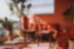 Sunlit Moroccan-style patio with red patterned cushions, wooden tables, potted plants, and ornate railing casting shadows.