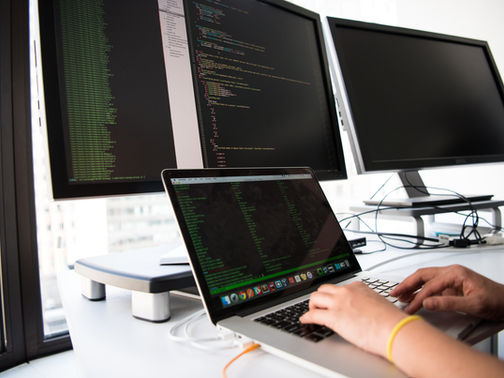 Person typing on laptop at a bright desk with three monitors showing code, a focused programmer in a modern offic