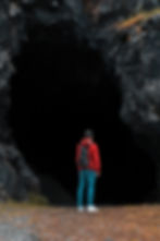 Young man with a bookbag on his back stands at the mouth of a large cave
