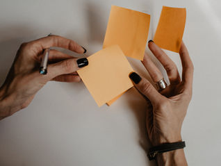 Hands with black nails holding orange sticky notes and a pen. Against a neutral background.