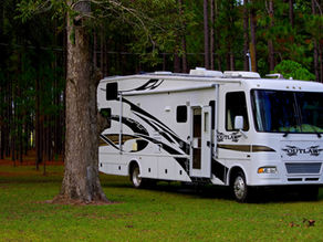 White RV marked "Outlaw" parked in a lush green forest. Tree trunks in the background. Calm and serene outdoors setting.