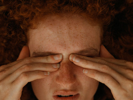 Person with curly red hair, freckles, and closed eyes, pressing fingers on eyelids. Wearing a knitted white sweater. Calm expression.
