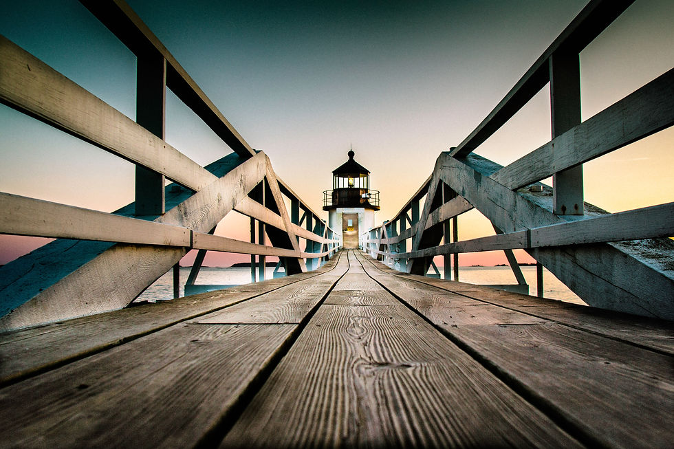 Wooden pier leads to a lighthouse at sunset. The sky is a gradient of pink, orange, and blue, creating a serene, peaceful mood.