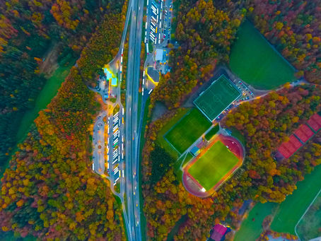 Aerial view of highways, parking lots, and sports fields amidst colorful autumn trees. Green and reddish fields on the right, vibrant foliage.