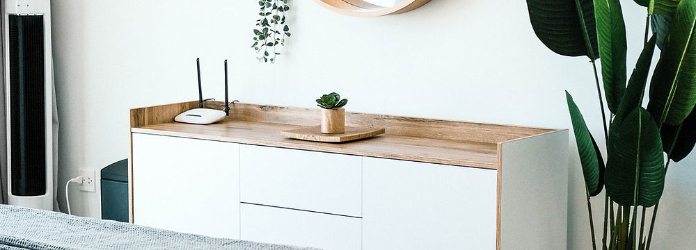 A clean and minimalist bedroom featuring a wooden bed, a white and wood dresser, and a large plant in a glass vase. A round wooden mirror hangs on the white wall above the dresser.