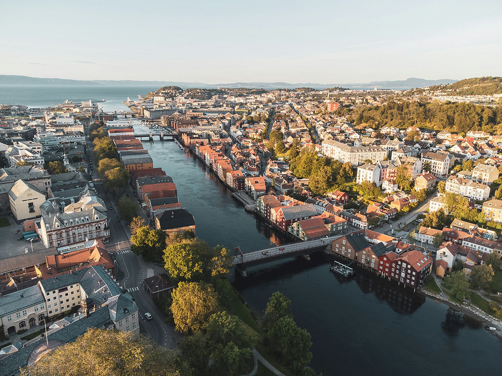 Luftaufnahme von Trondheim mit Fluss, Brücken, roten Dächern, Hafen und Meer im Hintergrund bei warmem Licht.
