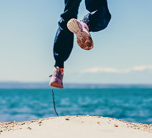 Mensch springt in die Luft, Beine und Turnschuhe, Meer und Strand. Image by Jim Makos