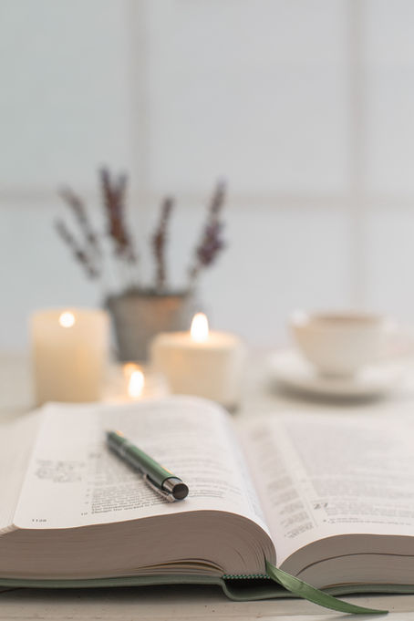 Open bible with a green pen on top, surrounded by lit candles. Lavender in the background and a cup on a saucer. Calm, cozy setting.