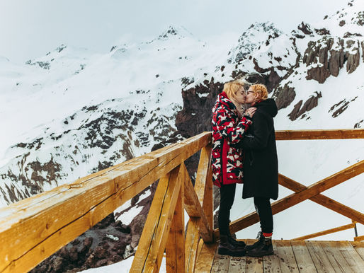 Two women kiss on a wooden mountain lookout, wearing winter coats, with snowy peaks and cliffs in the background.