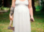 Person in white dress holds an axe and a jar outdoors. Arm tattoo visible. Green grass in background, conveying a rustic vibe.