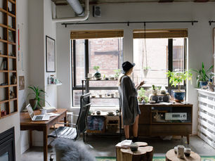 A woman in a hat tends to plants in a cozy room with large windows. A desk with a laptop and a shelf with decor are visible. Bright, modern vibe.
