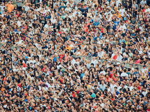 Dense crowd of diverse spectators at an outdoor event, many wearing lanyards and raising phones, packed behind barriers.
